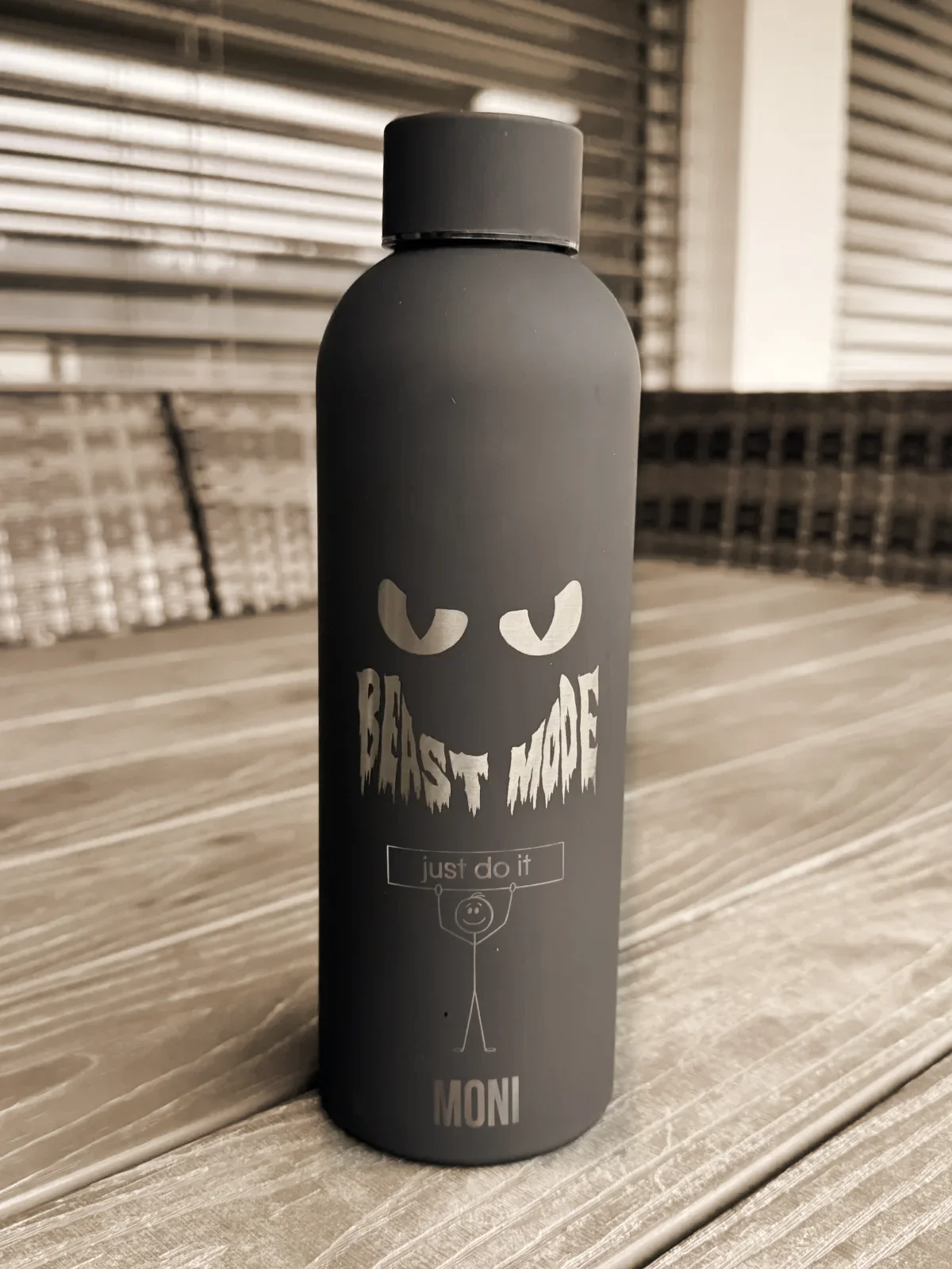 Mattschwarze Thermosflasche mit der Gravur Beast Mode, einem Strichmännchen mit Schild just do it und dem Namen Moni, auf einem Holztisch vor Lamellenstoren
