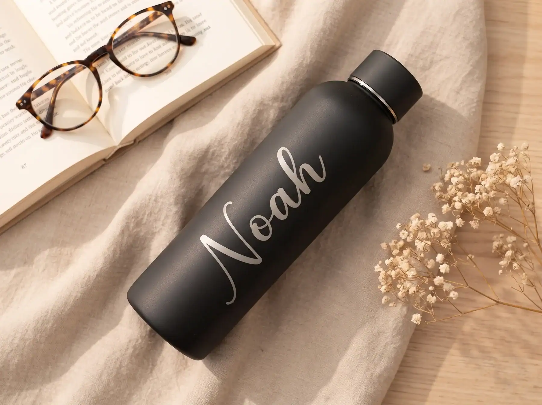 Schwarze Thermosflasche mit dem gravierten Namen Noah, liegend neben Buch und Brille